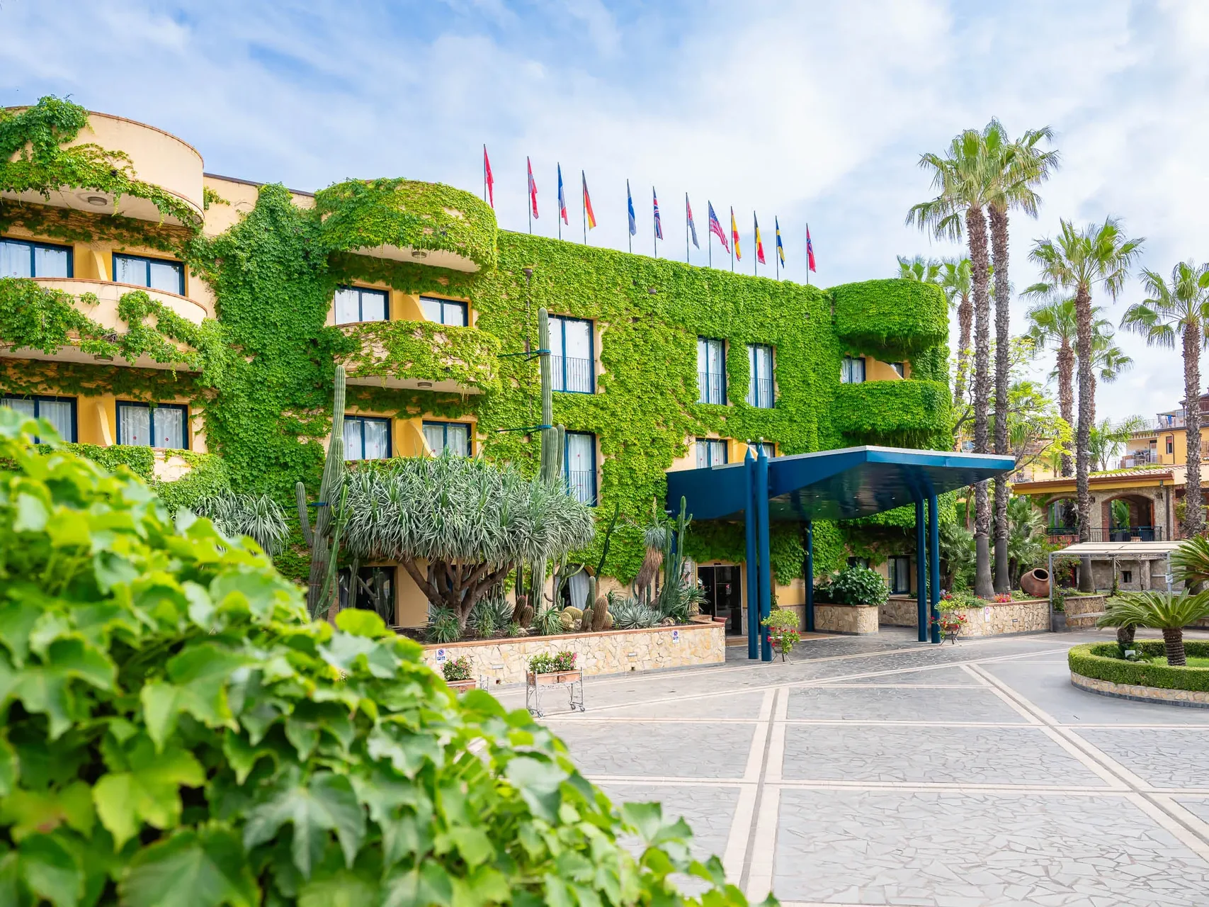 Facciata dell'hotel ricoperta di edera con bandiere internazionali, palme e piante grasse all'ingresso