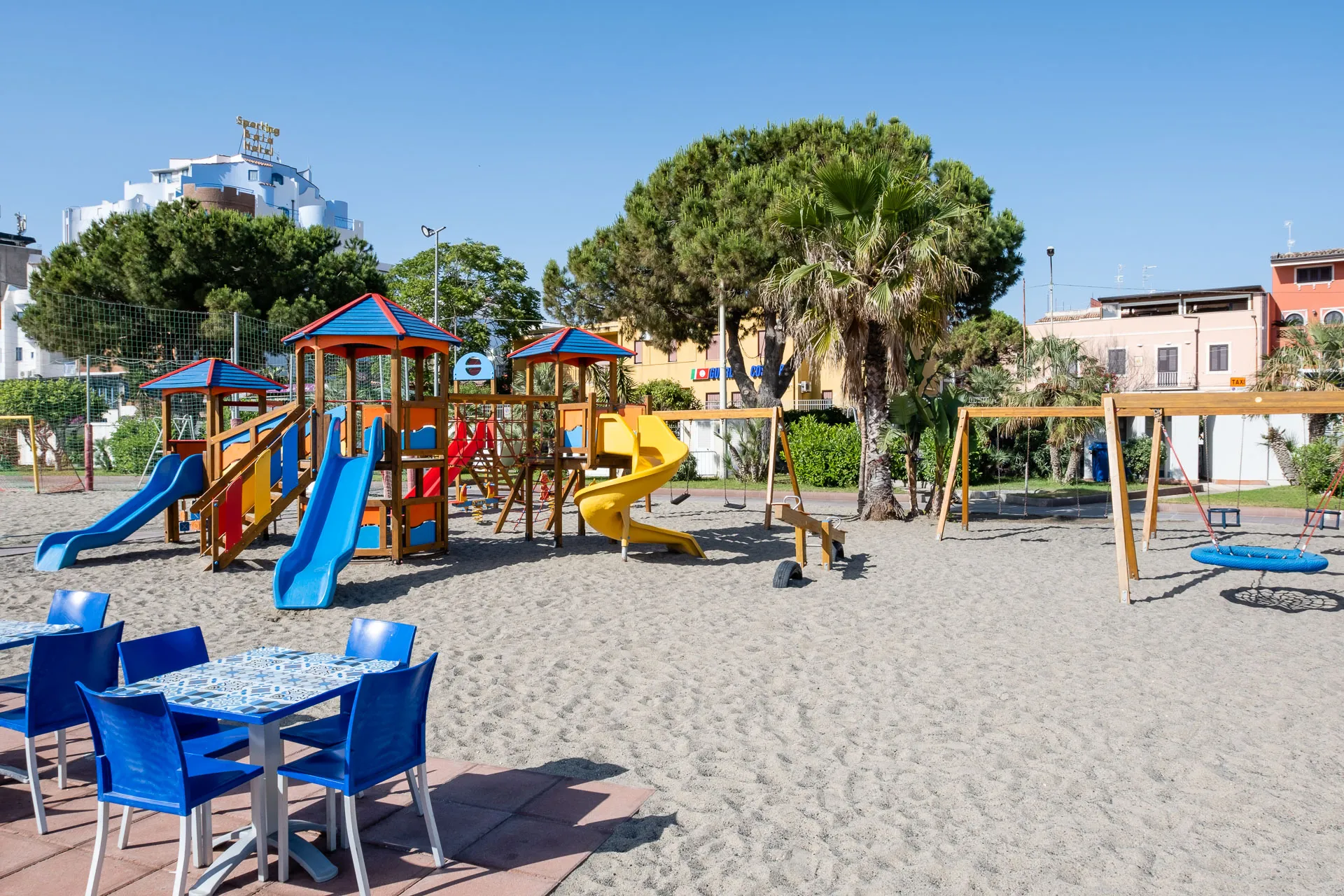 Area giochi per bambini con scivoli colorati, altalene e tavoli su terreno sabbioso