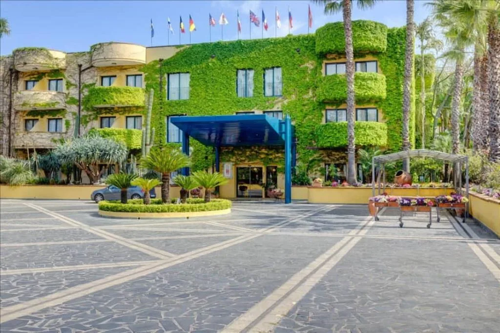 Ingresso dell'hotel con facciata ricoperta di edera verde, palme, bandiere e ampio piazzale