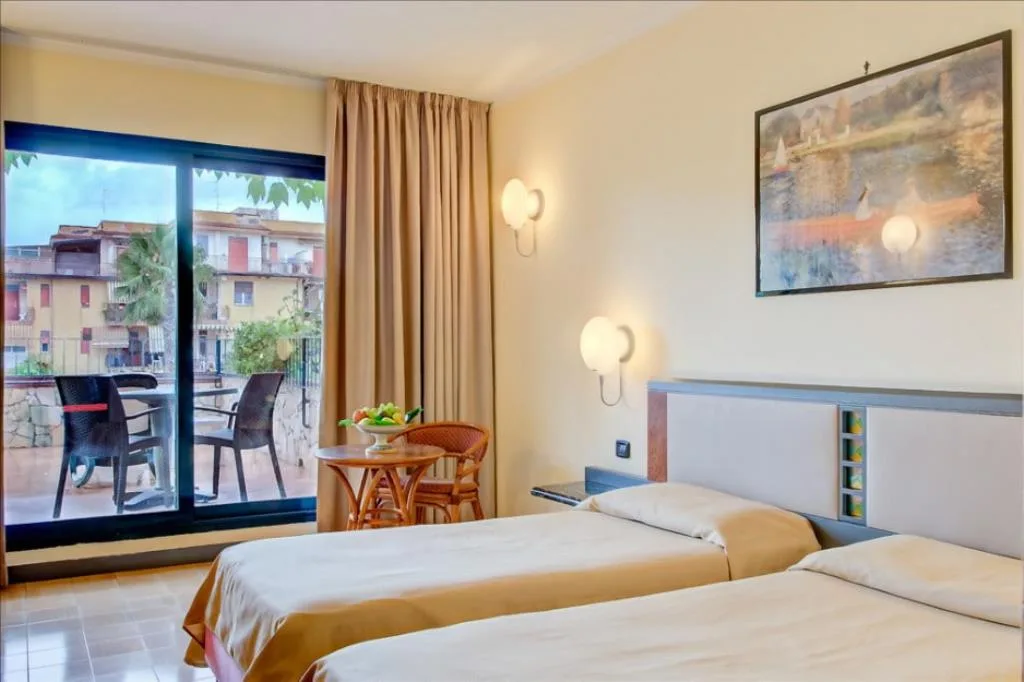 Camera con due letti, balcone affacciato su edifici e palme, tavolino con frutta e quadro decorativo