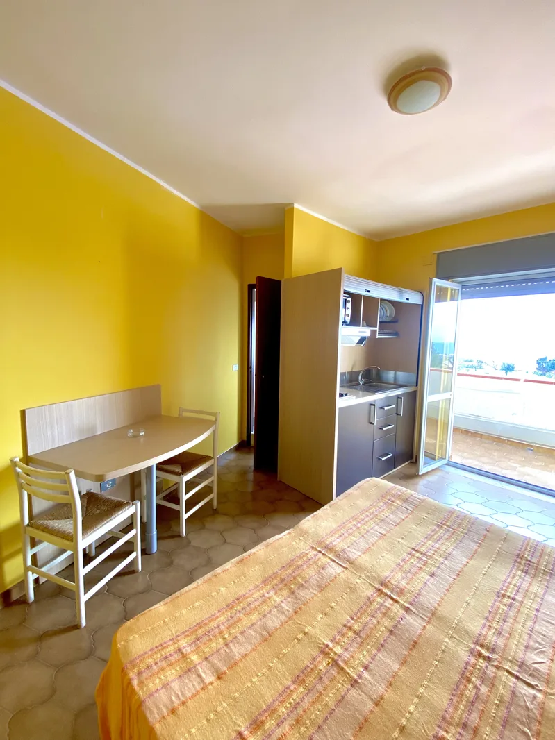 Camera con pareti gialle, letto matrimoniale, tavolo da pranzo e angolo cottura con accesso al balcone