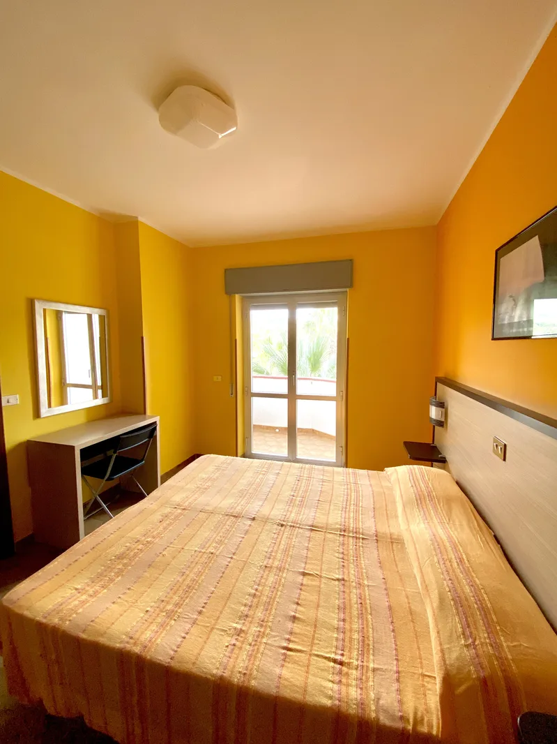 Camera da letto matrimoniale con pareti gialle, scrivania con specchio e porta-finestra che dà sul balcone