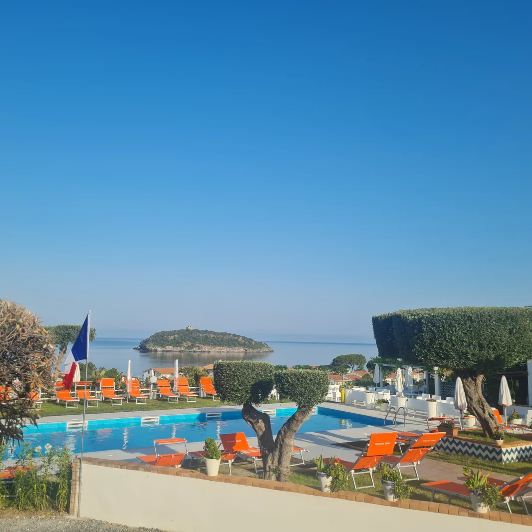 Piscina con lettini arancioni e ulivi affacciata sul mare e isolotto sotto cielo azzurro