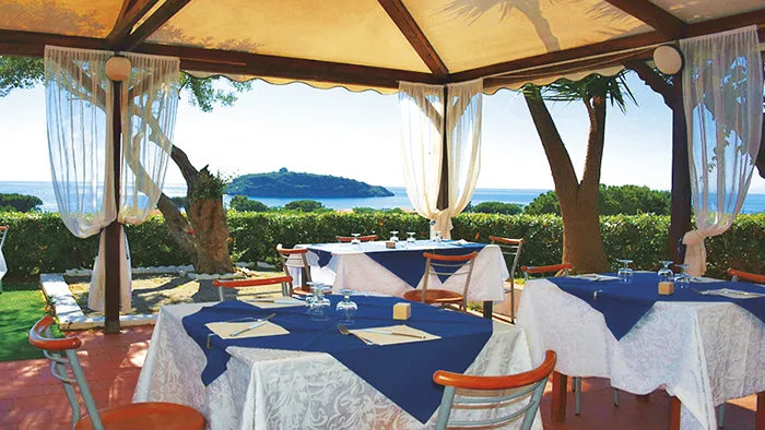 Ristorante esterno sotto gazebo con tavoli blu e tende bianche affacciato sul mare e isolotto