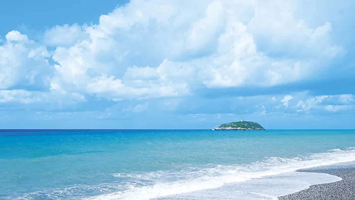 Spiaggia di ciottoli con mare azzurro, cielo nuvoloso e isolotto all'orizzonte