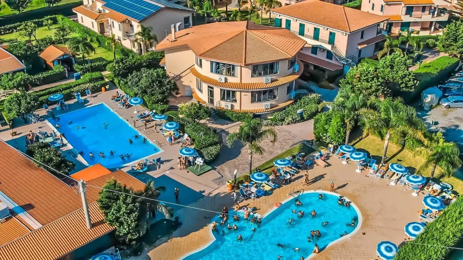 Veduta aerea del villaggio con piscine, ombrelloni e bagnanti tra gli edifici
