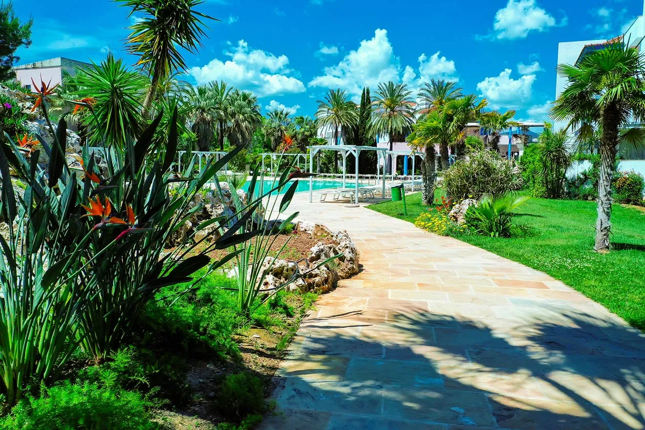 Vialetto in pietra tra giardini rigogliosi con palme e fiori tropicali verso la piscina