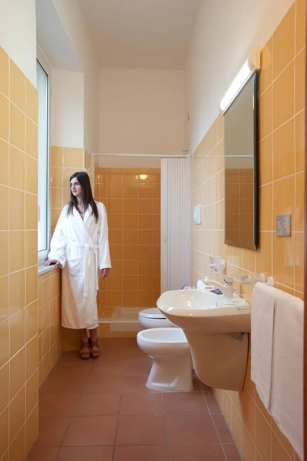 Bagno con piastrelle arancioni, lavabo, bidet, water e donna in accappatoio bianco accanto alla finestra