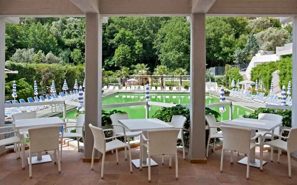Terrazza coperta con tavoli e sedie bianche affacciata sulla piscina circondata dal verde