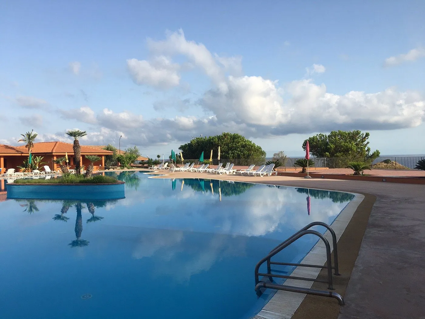 Piscina con isolotto di palme che si riflette nell'acqua e vista mare al tramonto