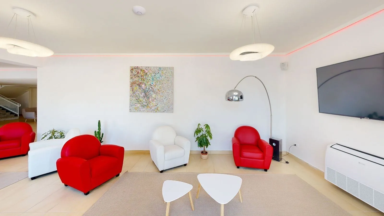 Sala lounge moderna con poltrone rosse e bianche, TV a parete e tavolini triangolari