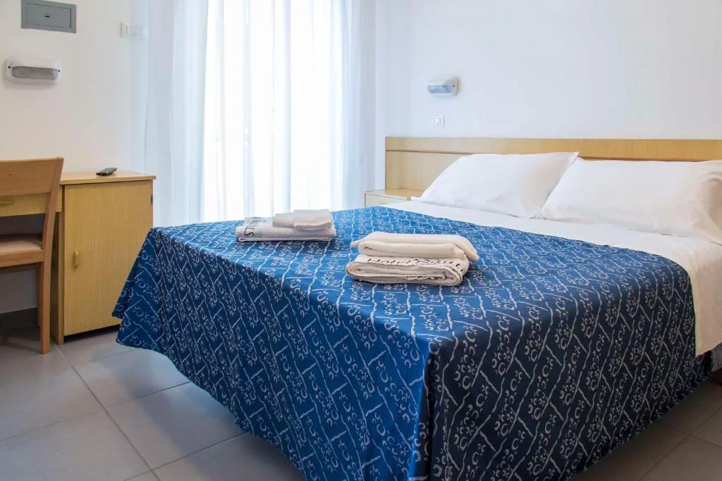 Camera con letto matrimoniale con copriletto blu, asciugamani, scrivania in legno e finestra luminosa