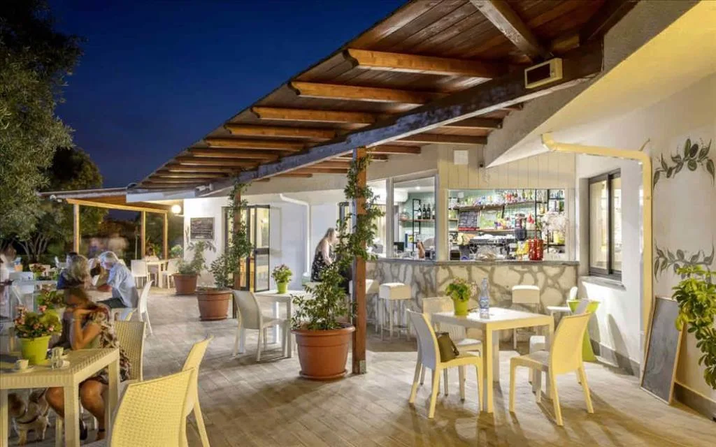 Terrazza bar all'aperto serale con tavoli, sedie gialle e tettoia in legno
