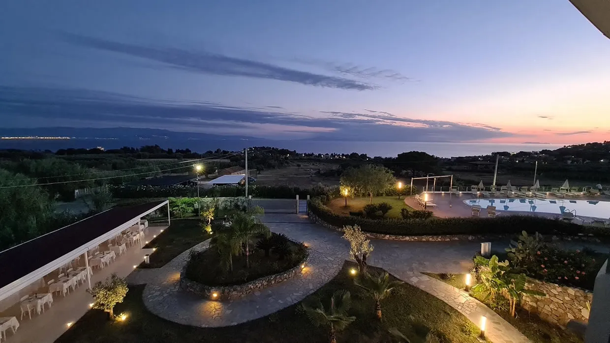 Vista serale del resort con giardini illuminati, piscina e tramonto sul mare