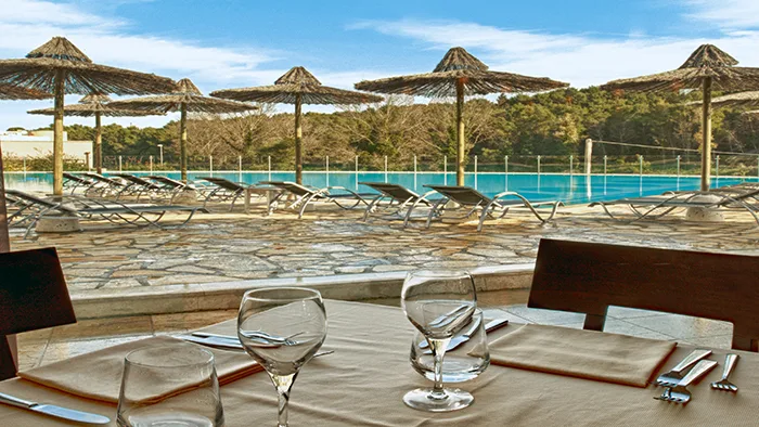 Tavolo del ristorante apparecchiato con vista sulla piscina e ombrelloni in paglia
