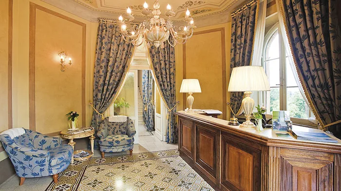 Elegante hall con lampadario, tende decorate, poltrone blu e reception in legno