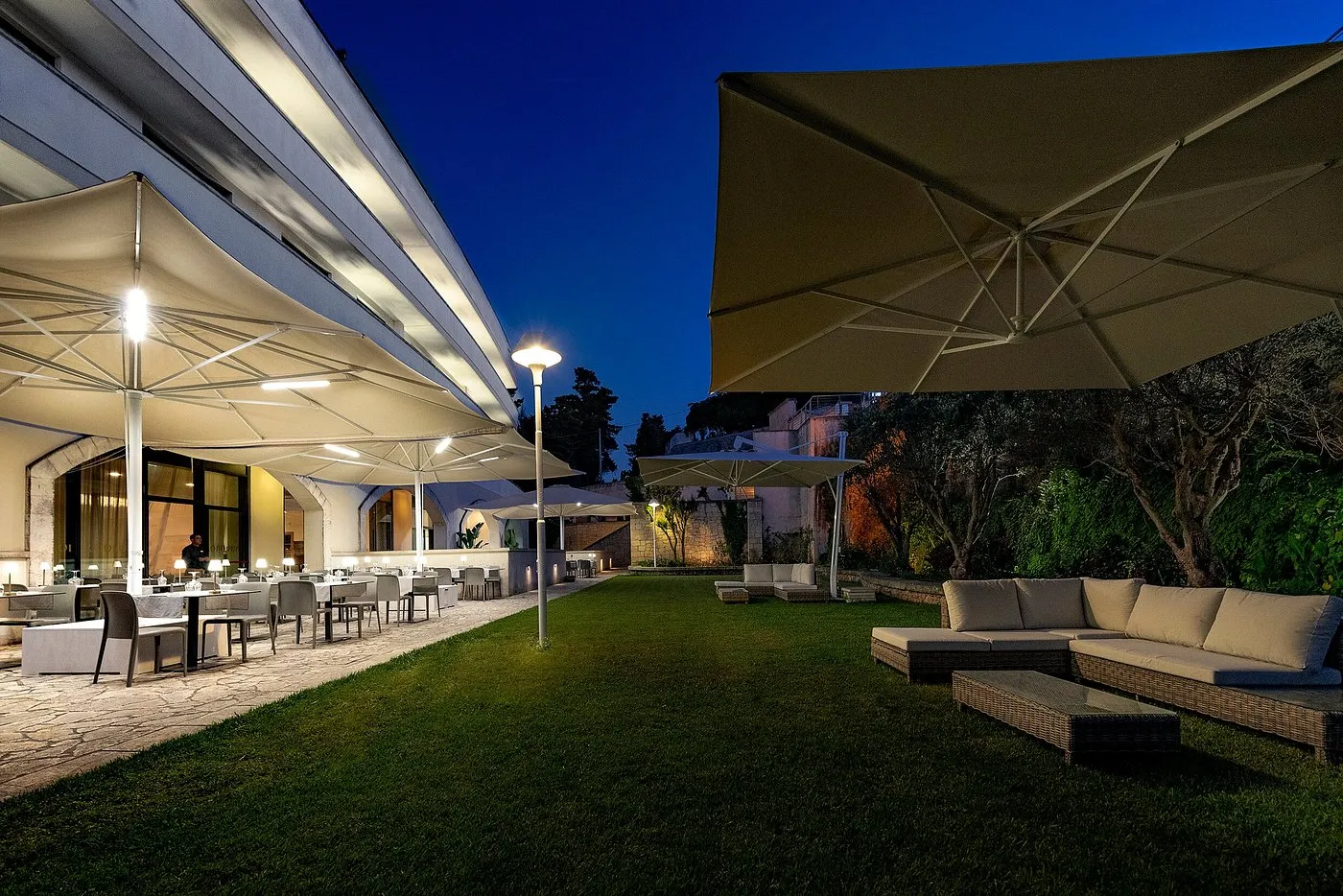 Terrazza esterna serale con tavoli del ristorante, ombrelloni illuminati e divani lounge sul prato