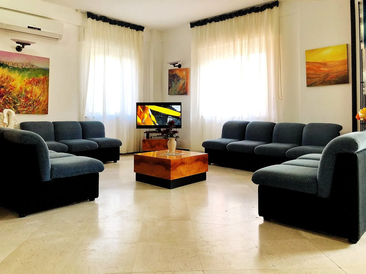 Hall con divani blu, tavolino in legno, televisore e quadri colorati alle pareti