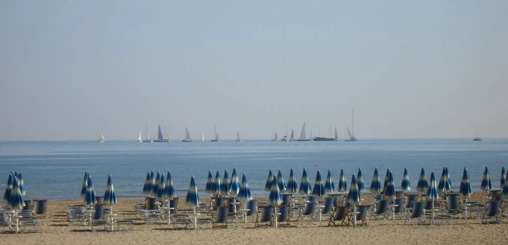 Spiaggia con ombrelloni chiusi e mare con barche a vela all'orizzonte