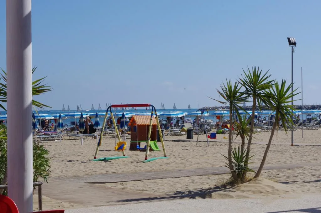 Spiaggia sabbiosa con giochi per bambini, ombrelloni e mare con barche a vela