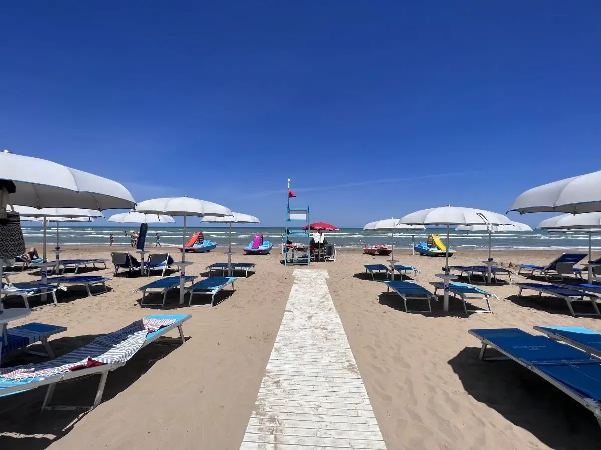 Spiaggia sabbiosa con passerella in legno, ombrelloni bianchi, lettini blu e pedalò vicino al mare