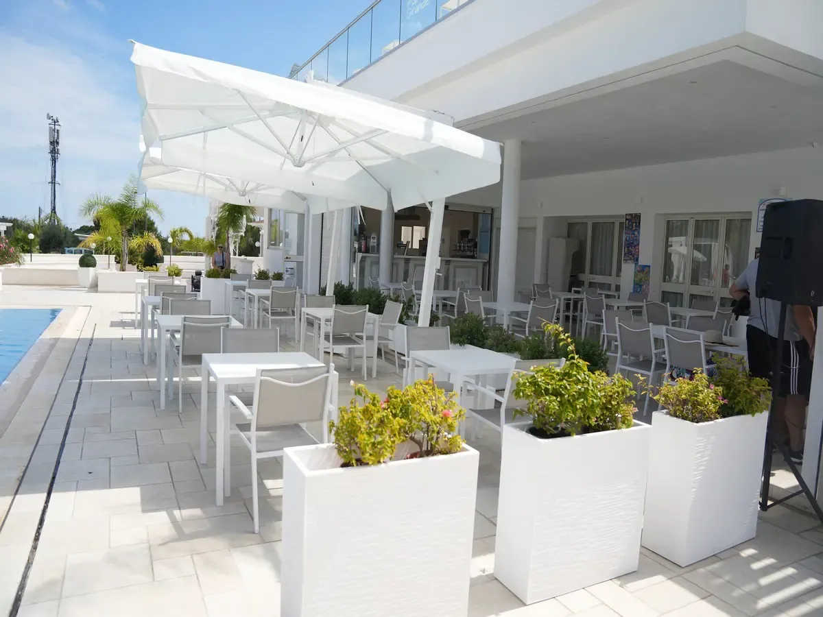 Terrazza bar moderna con tavoli e sedie bianche, fioriere e ombrellone accanto alla piscina