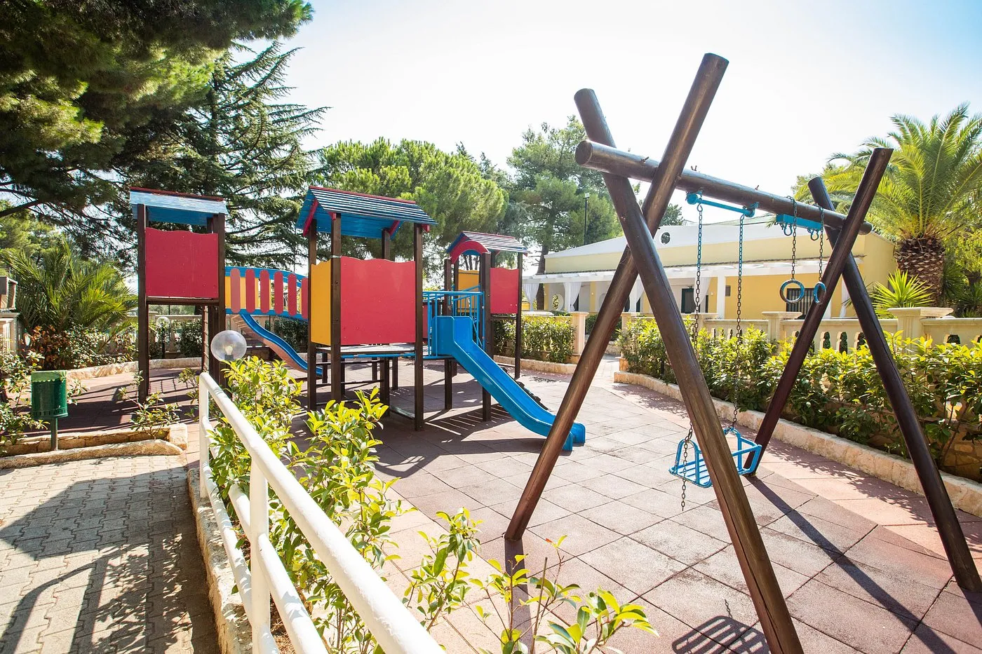 Area giochi per bambini con scivolo blu, altalene e struttura colorata circondata da pini