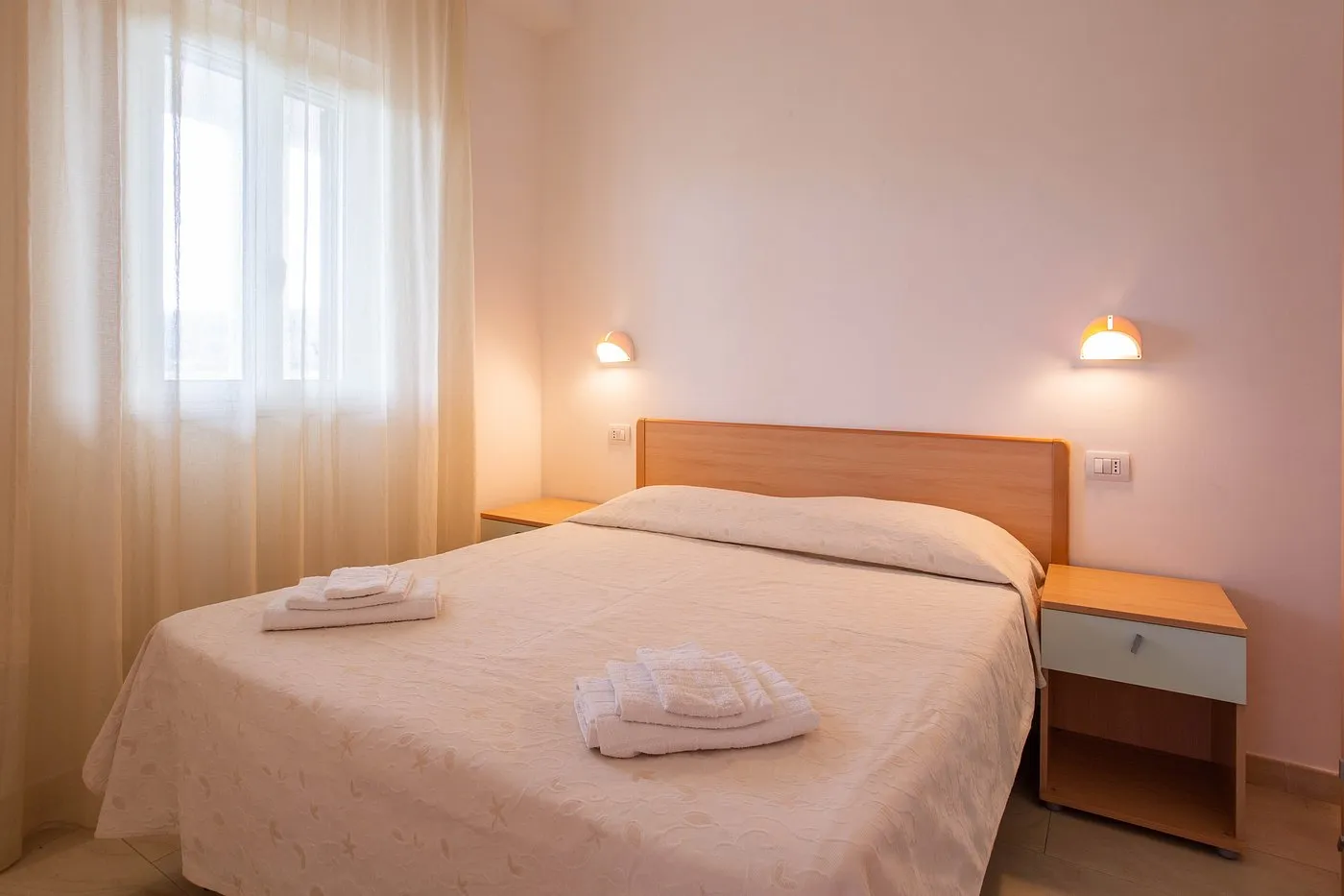 Camera da letto con letto matrimoniale, comodini in legno e asciugamani, illuminata da luce naturale