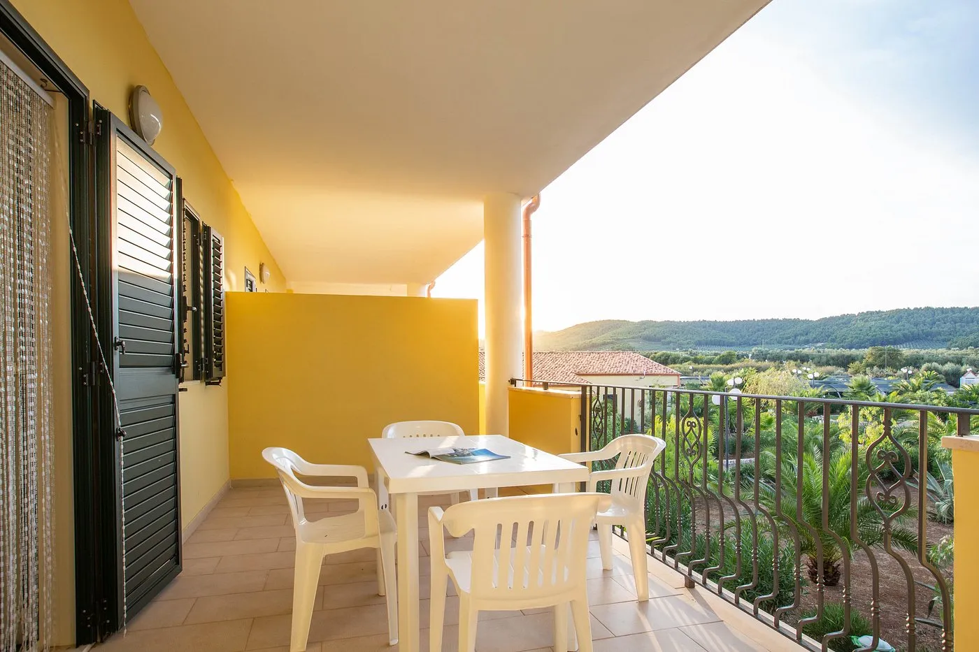 Balcone con tavolo e sedie bianche, vista sul giardino con palme e colline al tramonto