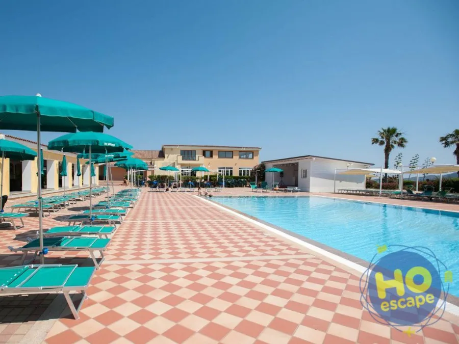 Piscina con pavimentazione a scacchi, ombrelloni verdi e lettini davanti all'edificio del resort