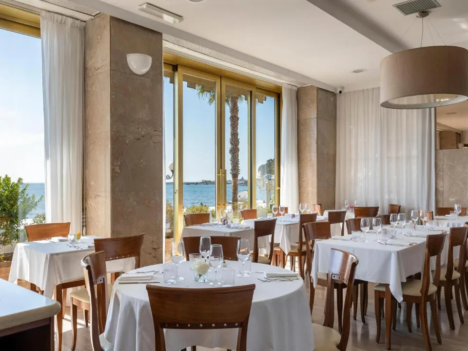 Sala ristorante con tavoli apparecchiati di bianco e ampie vetrate affacciate sul mare