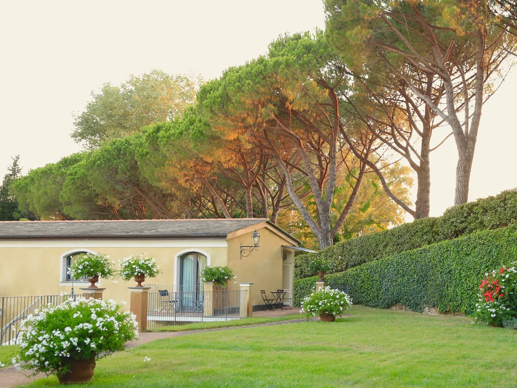 Esterno dell'hotel con edificio giallo, giardino fiorito e pini al tramonto
