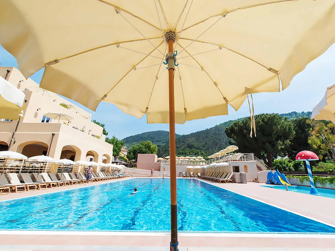 Piscina vista da sotto un ombrellone giallo con scivolo per bambini e colline sullo sfondo