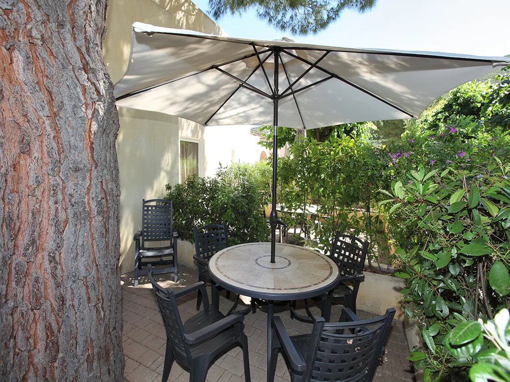 Patio esterno con tavolo rotondo, sedie nere e ombrellone accanto a un grande albero