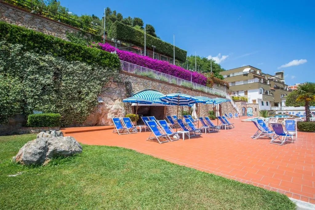 Area solarium con lettini e ombrelloni blu su pavimentazione, muro fiorito con bouganville e prato verde