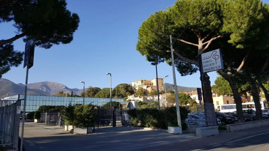Ingresso esterno del Riviera Palace Residence con insegna, alberi, parcheggio e colline sullo sfondo