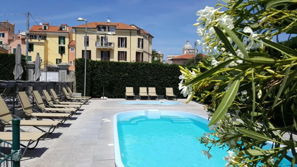 Piscina con lettini allineati, siepi e edifici colorati sullo sfondo con cespuglio fiorito bianco