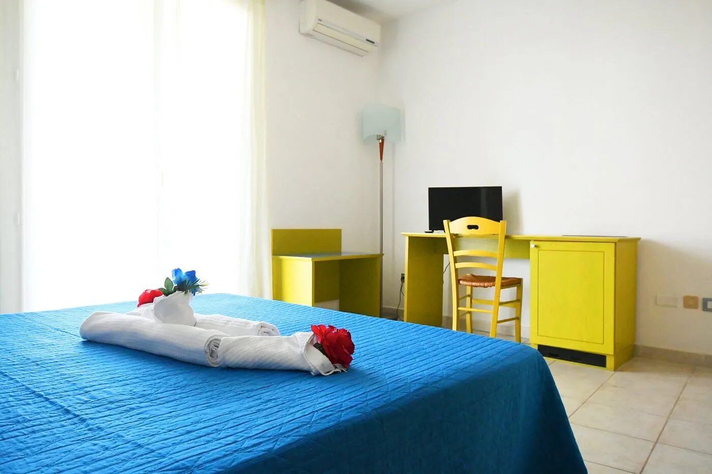 Camera da letto con copriletto azzurro, scrivania gialla e TV