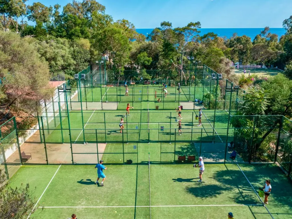 Campi da padel e tennis in erba con persone che giocano, circondati dalla vegetazione e dal mare