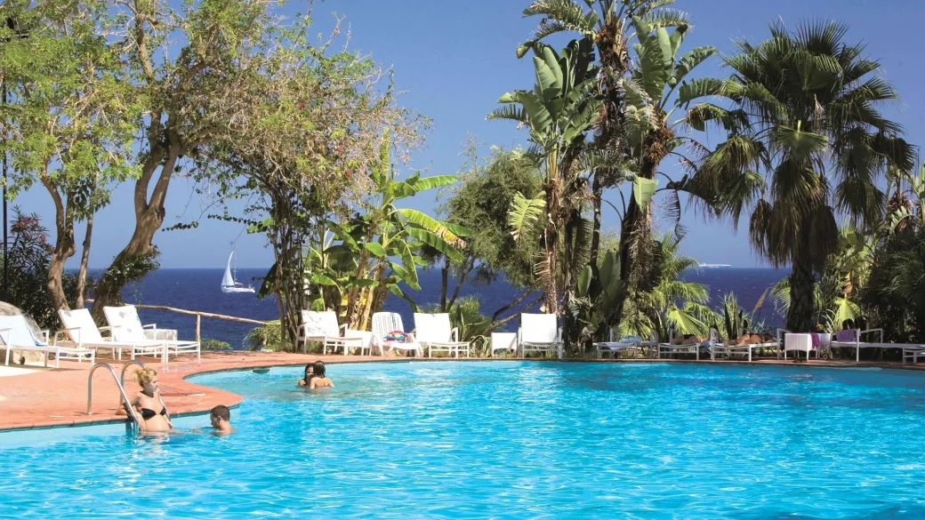 Piscina circondata da palme e vegetazione tropicale con vista sul mare e barca a vela