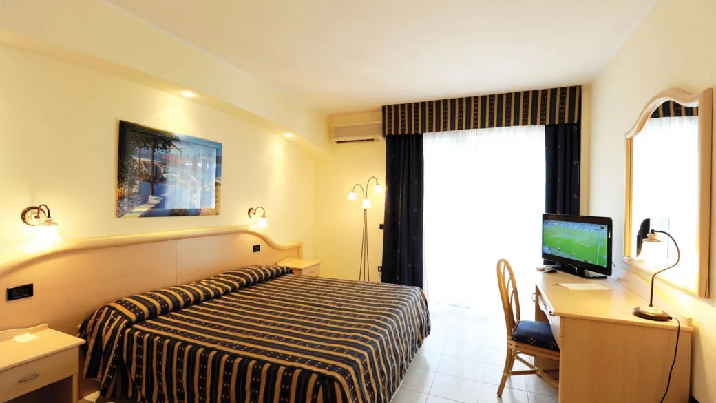 Camera con letto matrimoniale, copriletto a righe blu, scrivania, televisore e finestra luminosa