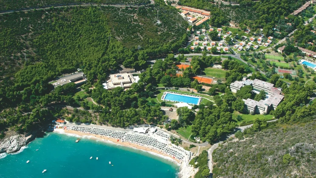 Veduta aerea del resort con piscina, campi da tennis e ville immerse nella pineta sopra la spiaggia