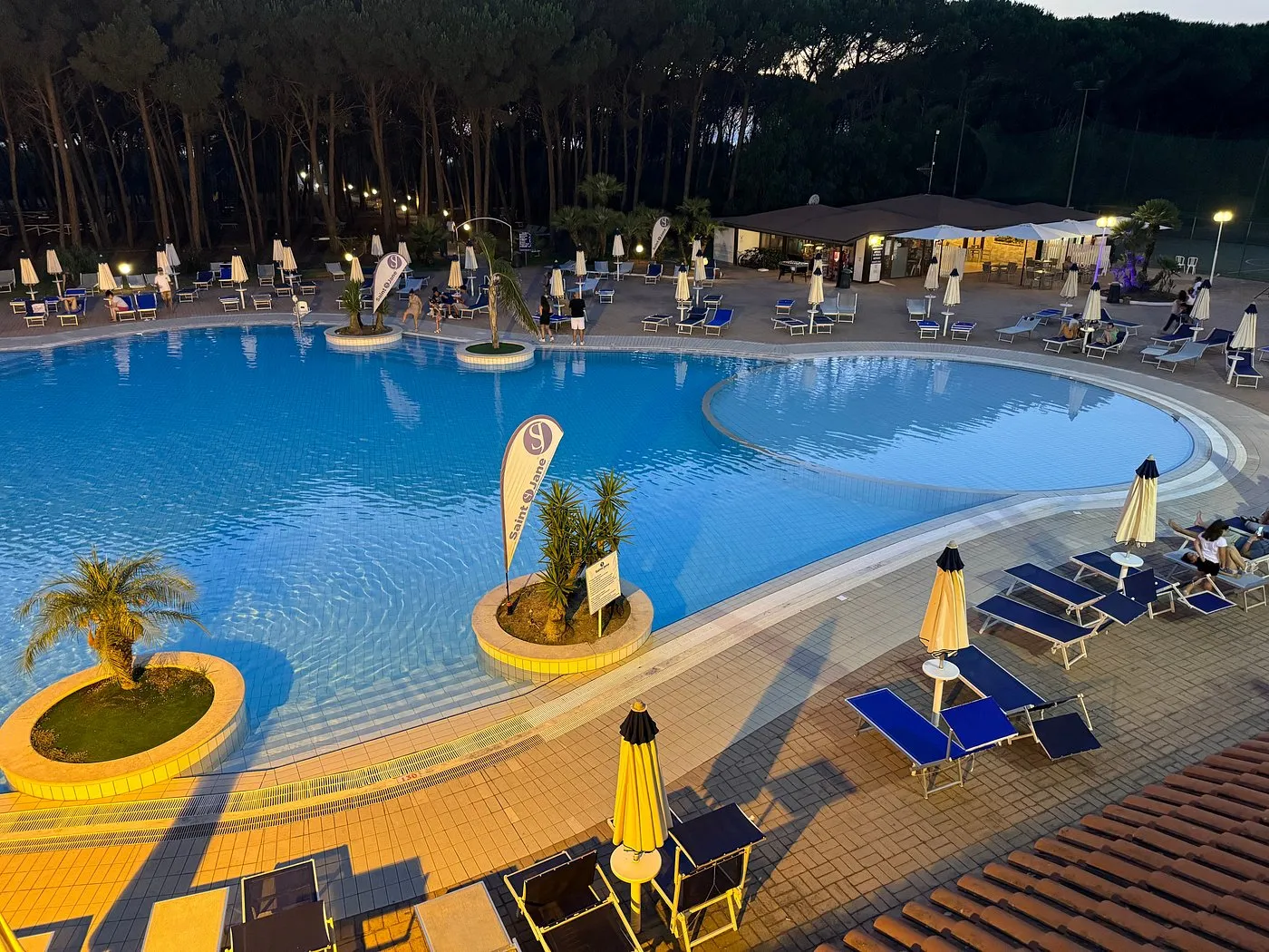 Piscina del resort al crepuscolo con lettini blu, ombrelloni e bar sullo sfondo