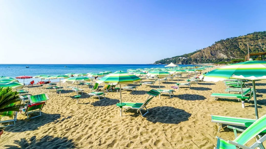 Spiaggia con ombrelloni e lettini verdi, mare azzurro e costa rocciosa