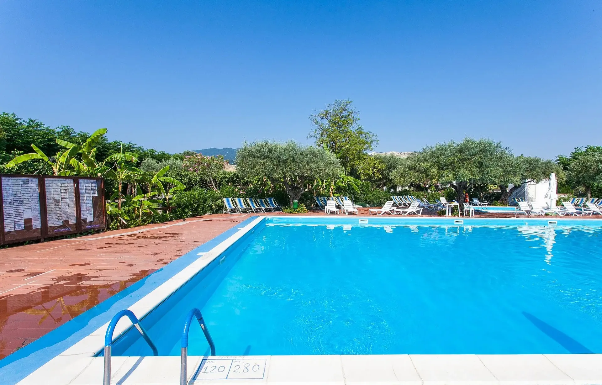 Piscina con indicazione della profondità e lettini sul bordo tra gli alberi