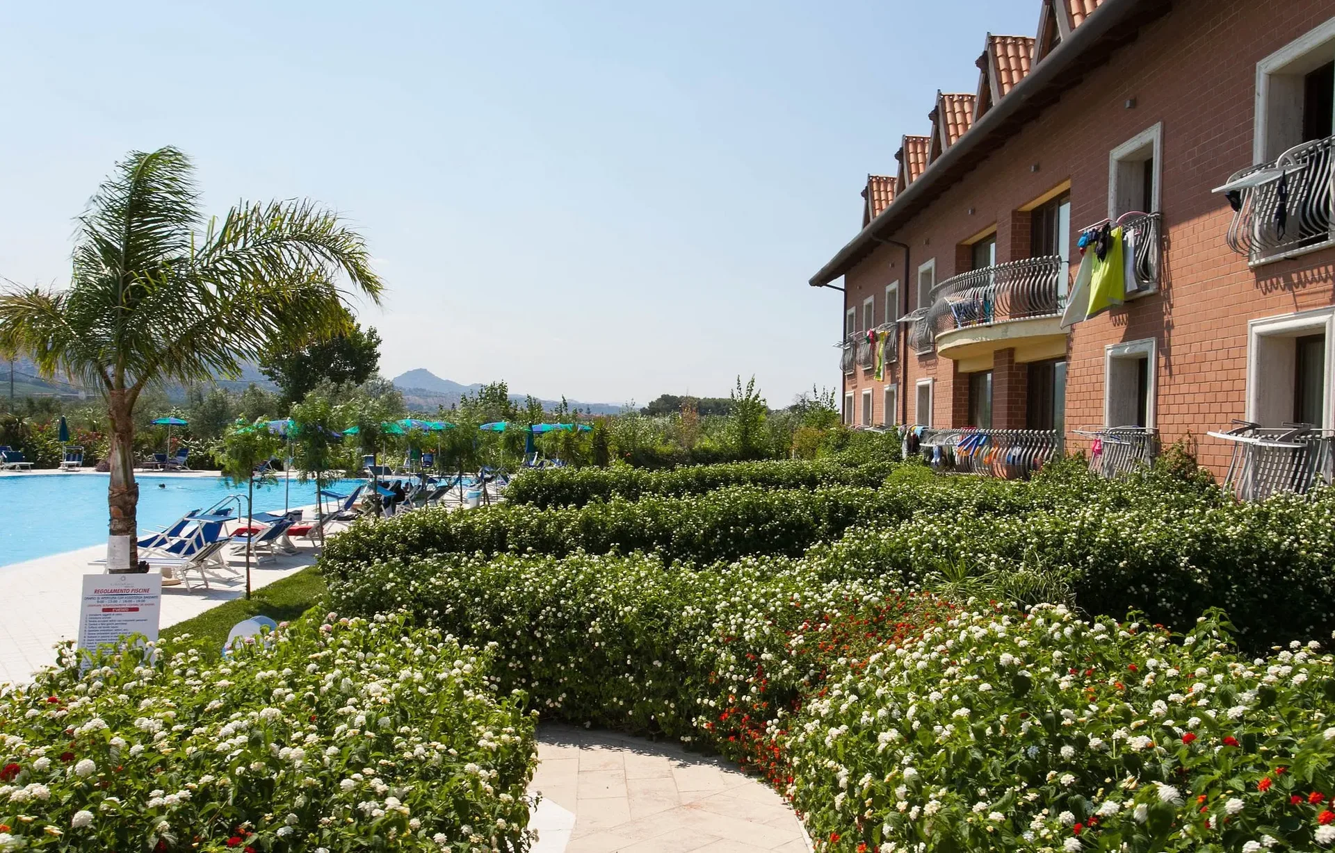 Giardino fiorito con siepi e palme lungo l'edificio del resort e la piscina