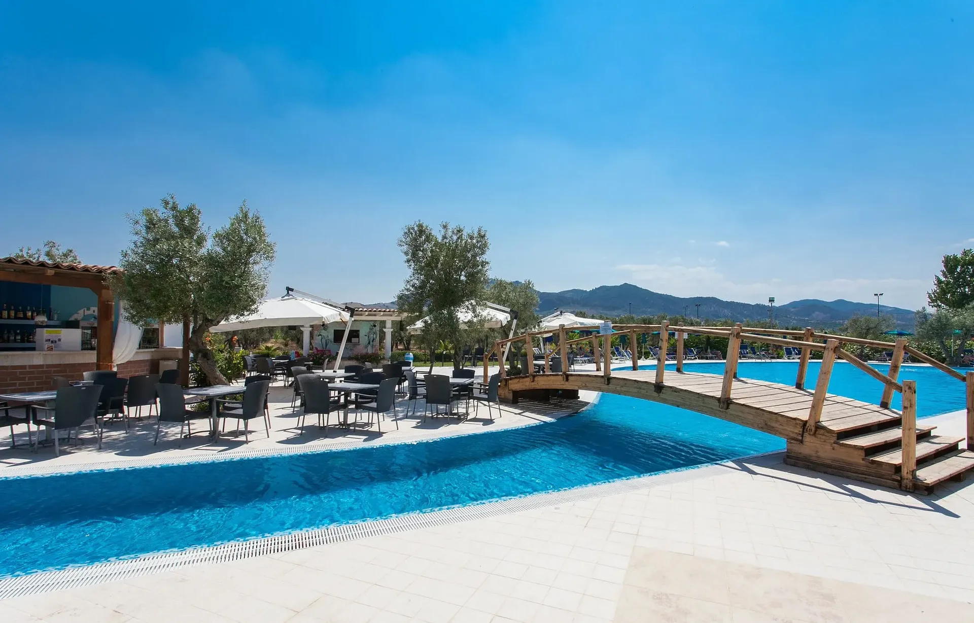Area bar a bordo piscina con tavoli, ombrelloni e ponte in legno tra gli ulivi
