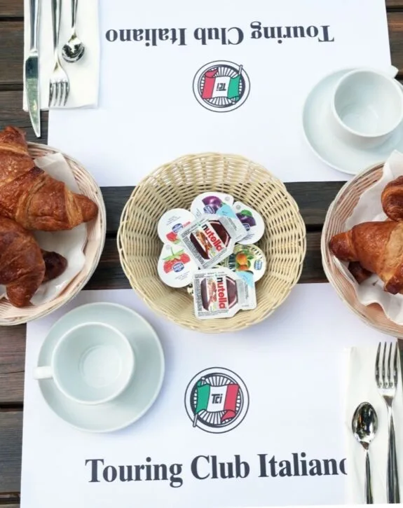 Colazione al Touring Club Italiano: croissant, burro, marmellate e tazza su un tavolo in legno.