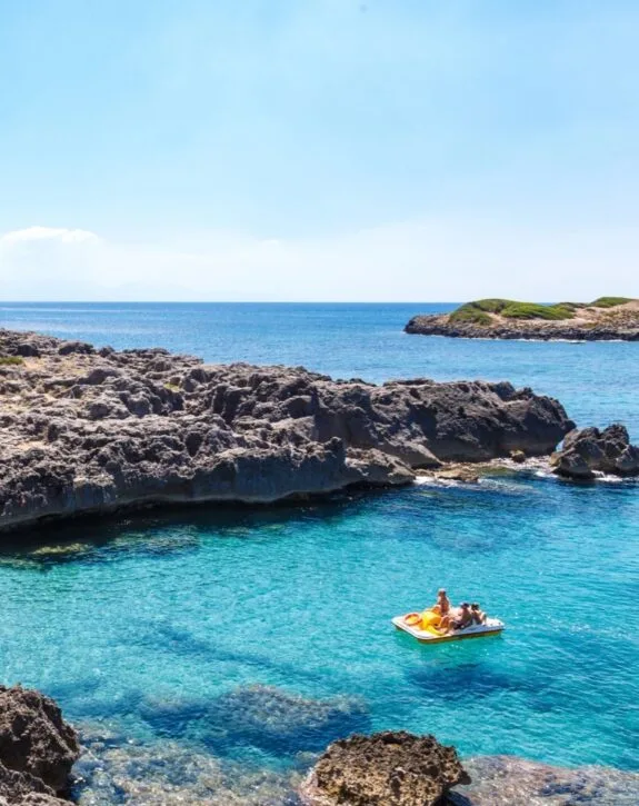 Due persone su un gonfiabile giallo nel mare cristallino, circondato da rocce e un’isola verde in lontananza.