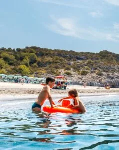 Due bambini giocano in acqua su un materassino arancione, con una spiaggia e colline verdi sullo sfondo.